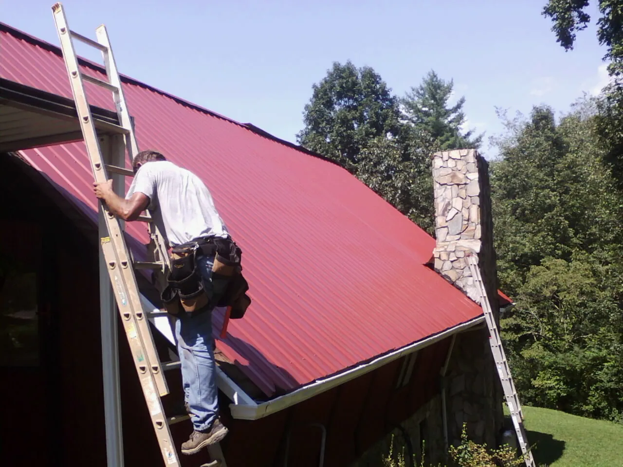 Roofer on a ladder beside a red metal roof and stone chimney — Soffit & Fascia Repair by Roofing Brothers in Superior